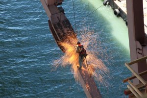 Engineer Martin Chandrawinata, on the new San Francisco-Oakland Bay Bridge, photographed an iron worker as he lance cut a section of the temporary truss on the self-anchored suspension structure. This truss carried the load of the bridge prior to the transfer to the cable.